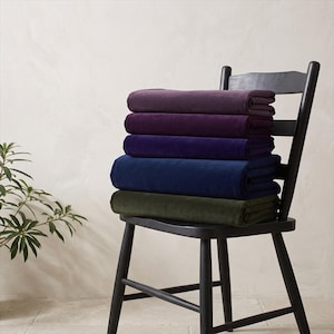 May include: A stack of five folded velvet blankets in shades of dark plum, deep purple, royal blue, navy blue, and olive green, resting on a black wooden ladder-back chair. The blankets have a soft, smooth texture and are neatly layered.