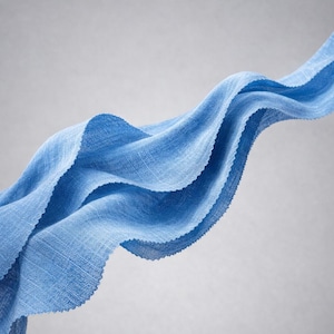 May include: A flowing, light blue fabric with a textured weave. The fabric is arranged in graceful, undulating waves against a soft gray background. The edges of the fabric are slightly frayed, adding to its delicate appearance.