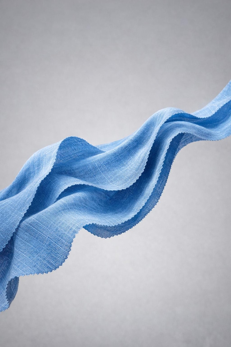 May include: A flowing, light blue fabric with a textured weave. The fabric is arranged in graceful, undulating waves against a soft gray background. The edges of the fabric are slightly frayed, adding to its delicate appearance.
