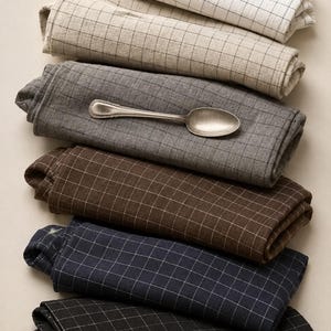 May include: A stack of six folded rectangular fabric napkins, each featuring a grid pattern. The colors include white, beige, gray, brown, navy blue, and black. A single silver-toned metal spoon rests horizontally on the gray napkin.