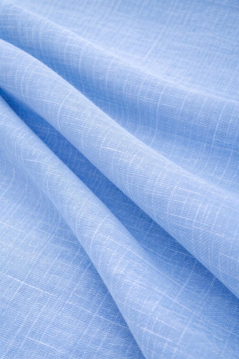 May include: Close-up of a light blue fabric with a subtle grid pattern. The fabric is draped, showing soft folds and textures. The color is a solid, pale blue, suitable for clothing or home decor.