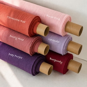 Pode incluir: Seis rolos de tecido em tubos de papelão, rotulados com o texto: Blush Pink, Terracotta Red, Dusty Rose, Lavender, Deep Purple e Burgundy. O tecido tem uma textura visível e está disposto em uma pilha, exibindo uma variedade de cores sólidas.