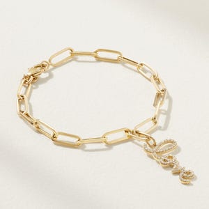 Könnte beinhalten: Goldfarbenes Kettengliederarmband mit einem 'Love'-Anhänger. Der Anhänger ist in einer Schreibschrift geschrieben und mit kleinen, klaren Edelsteinen besetzt. Das Armband wird auf einer weißen Oberfläche präsentiert.