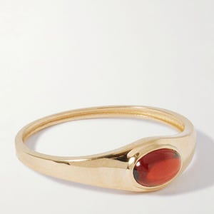 May include: A gold ring with a polished finish, featuring an oval-shaped, reddish-brown gemstone. The ring's band is smooth and slightly rounded, with the gemstone set in a bezel setting.