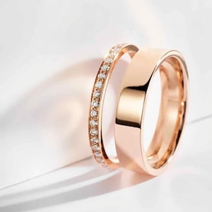 May include: Two rose gold wedding rings. One ring is a simple band, and the other is a band with a row of small, clear gemstones. The rings are displayed on a white background.