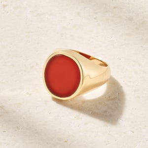 Può includere: Un anello con sigillo dorato con una grande pietra rossa ovale. L'anello ha una superficie lucida e riflettente e proietta un'ombra sulla superficie chiara su cui poggia. Un gioiello classico.