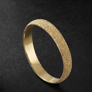 May include: A gold bangle bracelet with a textured, sandblasted exterior and a smooth, polished interior. The bracelet is a solid band of gold, showcasing a contrast between the textured outer surface and the shiny inner surface. The jewelry is set against a dark background.
