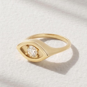 May include: A gold-colored ring with an eye-shaped design. The ring features a central, round, clear gemstone. The band and the eye-shaped setting are smooth and polished. The ring is set against a white background.