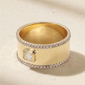 May include: A wide gold ring with a rectangular, clear gemstone set in a gold bezel. The ring is encircled by a row of small, clear gemstones. The band is approximately 0.5 inches wide.