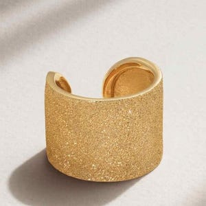 May include: A gold-colored ring with a textured, glittery surface. The ring has a wide band that curves around, leaving an opening. The ring is likely made of metal and is designed to be worn on a finger.