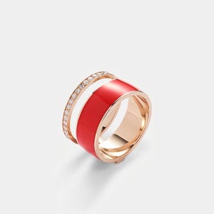 May include: A rose gold ring with a wide, glossy red enamel band and a thinner band encrusted with small, clear gemstones. The ring is set against a white background, highlighting its design.