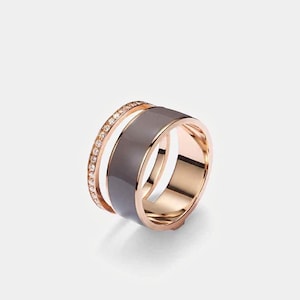 May include: A rose gold ring with a wide, dark gray band and a thinner band encrusted with small, clear gemstones. The ring has a modern, geometric design and a polished finish.