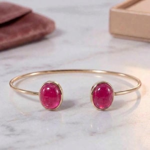 Könnte beinhalten: Ein goldfarbenes, offenes Armreif-Armband mit zwei ovalen, leuchtend rosa Edelsteinen. Die Edelsteine sind in einer Zargenfassung gefasst. Das Armband wird auf einer hellen Oberfläche präsentiert.