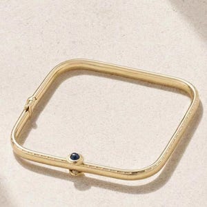 Könnte beinhalten: Ein goldfarbenes quadratisches Armreif-Armband mit einem kleinen, dunkelblauen Edelstein. Das Armband hat einen Klappverschluss und ein zartes, eingraviertes Muster an den Kanten. Das Design ist schlicht und elegant.