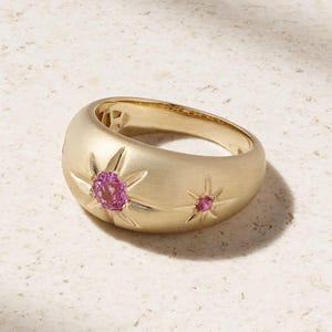 May include: A gold-colored ring with a domed design. The ring features two starburst designs, each with a pink gemstone at the center. The ring is set against a neutral background, highlighting the jewelry's details.