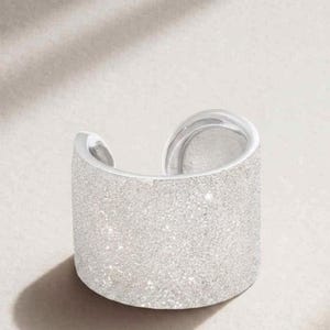 May include: A silver cuff bracelet with a textured, glittery surface. The open design allows for easy wear. The jewelry piece is a statement accessory.