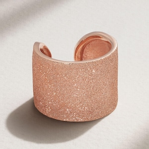 May include: A rose gold cuff ring with a textured, glittery surface. The open design allows for easy wear. The ring is a wide band, suitable for a statement piece.