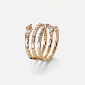May include: A gold-toned ring with a spiral design. The ring features multiple bands adorned with rectangular and round-cut clear gemstones. The gemstones are set along the bands, creating a sparkling effect. The ring is displayed against a white background.
