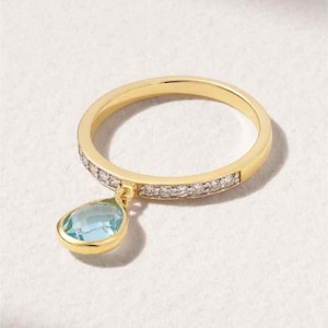 May include: A gold ring with a band encrusted with small, clear stones. A teardrop-shaped, light blue gemstone hangs from the ring. The ring is set against a white background.