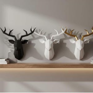May include: Three geometric deer head wall decor pieces. One is black, one is white, and the third is white with gold antlers. They are mounted on a light gray wall above a wooden shelf. The deer heads are a modern, minimalist design.