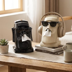 May include: Two ceramic tissue box holders shaped like Moai statues, one black and one cream-coloured, each wearing sunglasses. The cream-coloured statue has headphones. Both statues have tissue coming out of their mouths. They sit on a wooden table.