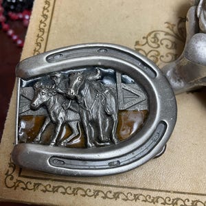 May include: A silver belt buckle shaped like a horseshoe with a raised design of three horses and riders. The buckle has a brown and gold enamel background.
