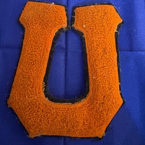 May include: Orange felt letter "U" with a black border, on a blue fabric background. The letter has a textured, fuzzy appearance, suggesting a vintage or collegiate style. The blue fabric has vertical folds.