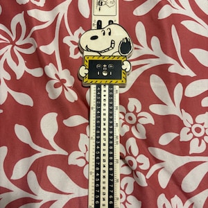 May include: A vintage Snoopy ruler featuring Snoopy holding a yellow and black sign. The ruler is marked with inch measurements up to 12 inches, and has Snoopy and other Peanuts characters at the top. The ruler is cream-colored with black markings.