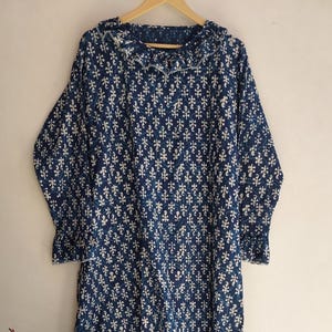 May include: A long, flowing dress in a dark blue hue, adorned with a repeating white floral pattern. The dress features long sleeves, a ruffled collar, and a tiered hemline. The garment is displayed on a wooden hanger.