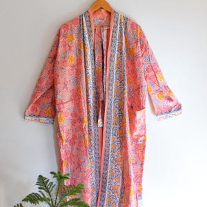 Puede incluir: Una bata larga y abierta con estampado floral. La bata es principalmente de color melocotón con flores amarillas y rosas, y detalles azules y blancos. Tiene mangas largas, bolsillos y un lazo con borla. La bata está colgada en una percha de madera.