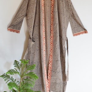 May include: A long-sleeved robe featuring a dark brown and white herringbone pattern throughout. The garment includes a contrasting orange and white decorative border along the front opening and sleeve cuffs, with a matching fabric tie at the waist.