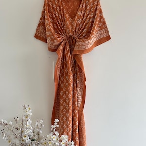May include: An orange kaftan dress with a V-neck and short sleeves, hanging on a wooden hanger. The dress features a white floral pattern and a tie at the waist. The dress is approximately 55 inches long.
