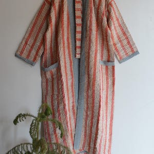May include: A long, open-front robe with a vertical striped pattern in coral and cream. The robe features grey trim on the sleeves, pockets, and collar. The robe is made of a quilted fabric.