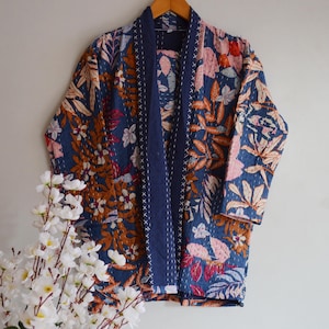 Puede incluir: Chaqueta acolchada con estampado floral en tonos azules, marrones y rosas. La chaqueta tiene un ribete azul marino y está colgada de una percha de madera. El diseño presenta una variedad de hojas y flores.