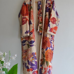 Könnte beinhalten: Eine lange, cremefarbene Robe mit einem lebendigen Blumenmuster in Rot-, Orange-, Lila- und Blautönen. Die Robe hängt an einem Holzkleiderbügel und zeigt ihre offene Front und langen Ärmel. Der Stoff scheint gesteppt zu sein.