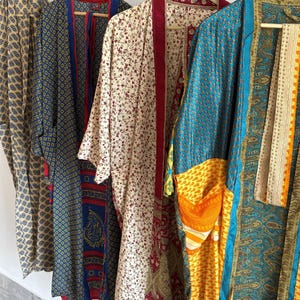 Puede incluir: Una colección de kimonos estampados y coloridos colgados de perchas de madera. Los kimonos presentan varios diseños, incluyendo estampados florales, paisley y geométricos en tonos azules, amarillos, rojos y crema. Las prendas están hechas de tela ligera.