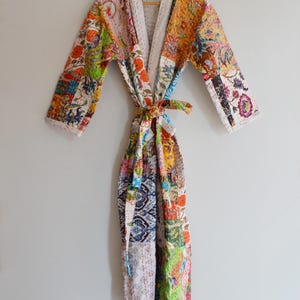 Könnte beinhalten: Bunter Patchwork-Bademantel mit floralem Muster. Der Bademantel hat einen Bindegürtel und weiße Verzierungen. Er besteht aus Stoffflicken in Orange, Grün, Rosa, Blau und Gelb. Der Bademantel hängt an einem Holzkleiderbügel.