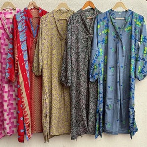 Puede incluir: Cinco vestidos largos de manga larga, colgados de perchas de madera, presentan diversos estampados y colores. Se distinguen estampados florales rosas, rojos, amarillos, grises y azules. Cada vestido es único.