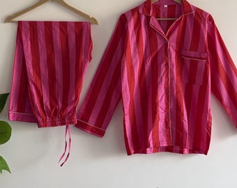 May include: A two-piece pajama set with a red and pink vertical stripe pattern. The long-sleeved button-down shirt has a chest pocket and white trim. The matching pants have a drawstring waist. The set is hanging on a wooden hanger.