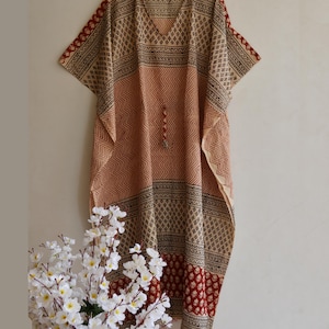 Könnte beinhalten: Ein beigefarbenes und rot gemustertes Kaftankleid mit V-Ausschnitt und kurzen Ärmeln. Das Kleid hat geometrische und florale Muster mit einem Zickzackmuster auf der Vorderseite. Das Kleid hängt an einem Holzkleiderbügel.