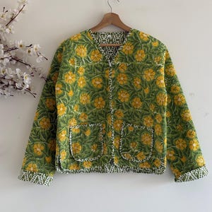 Puede incluir: Chaqueta acolchada verde con estampado floral amarillo y dos bolsillos delanteros. La chaqueta tiene cuello en V y es reversible, con forro estampado en blanco y verde. Cuelga de una percha de madera.