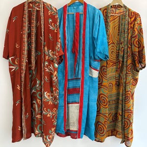 May include: Three colorful kimono robes hanging on wooden hangers. The robes feature various patterns and colors, including rust with floral designs, turquoise with red stripes, and gold with swirling patterns. Each robe has a unique design and texture.