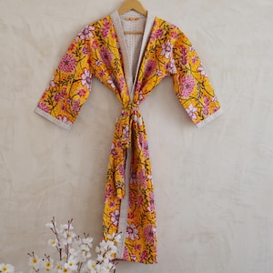 May include: A yellow floral patterned robe with pink and white flowers, hanging on a wooden hanger. The robe has a tie closure and a light-colored trim. The robe is made of a soft, quilted fabric.
