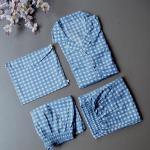 Può includere: Un completo pigiama a quadretti blu e bianchi, composto da una camicia abbottonata, pantaloni, pantaloncini e una borsa abbinata. Il pigiama ha bordini bianchi ed è disposto su una superficie grigia. Fiori bianchi sono nell'angolo in alto a sinistra.