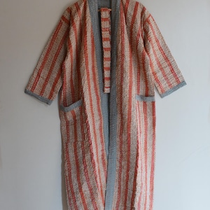 May include: A long, open-front robe with a striped pattern in shades of orange, cream, and gray. The robe has a shawl collar, pockets, and gray trim along the edges. It is hanging on a wooden hanger.