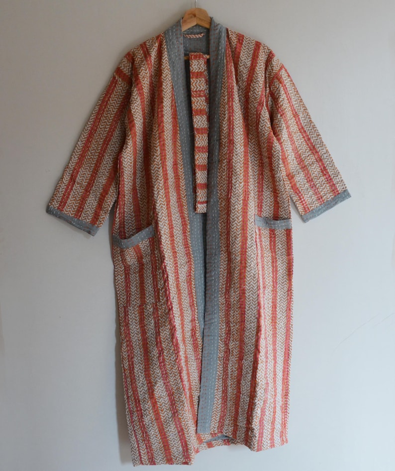 May include: A long, open-front robe with a striped pattern in shades of orange, cream, and gray. The robe has a shawl collar, pockets, and gray trim along the edges. It is hanging on a wooden hanger.