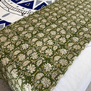 May include: A green and cream floral patterned quilt or throw. The fabric features a repeating design of stylized flowers and leaves. The quilt is folded over a white surface, with a blue and white patterned bedspread visible in the background.