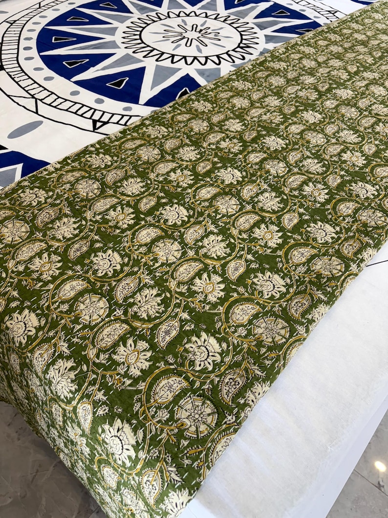 May include: A green and cream floral patterned quilt or throw. The fabric features a repeating design of stylized flowers and leaves. The quilt is folded over a white surface, with a blue and white patterned bedspread visible in the background.