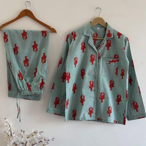 May include: A light blue pajama set with a red lobster pattern. The set includes a button-down shirt with a chest pocket and pants with a drawstring. The shirt and pants are hanging on wooden hangers.