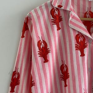 Puede incluir: Camiseta de pijama a rayas rosas y blancas con un estampado de langosta roja. La camiseta de manga larga tiene cuello y se abrocha por delante. El pijama est&aacute; colgado de una percha de madera.